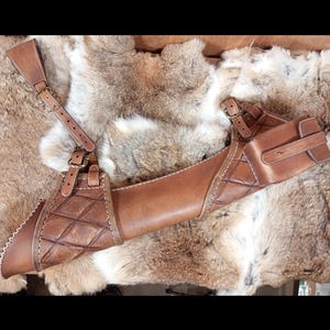 May include: A brown leather archery quiver with a shoulder strap and a small pouch. The quiver has a diamond pattern and white stitching. The leather is a medium brown color.
