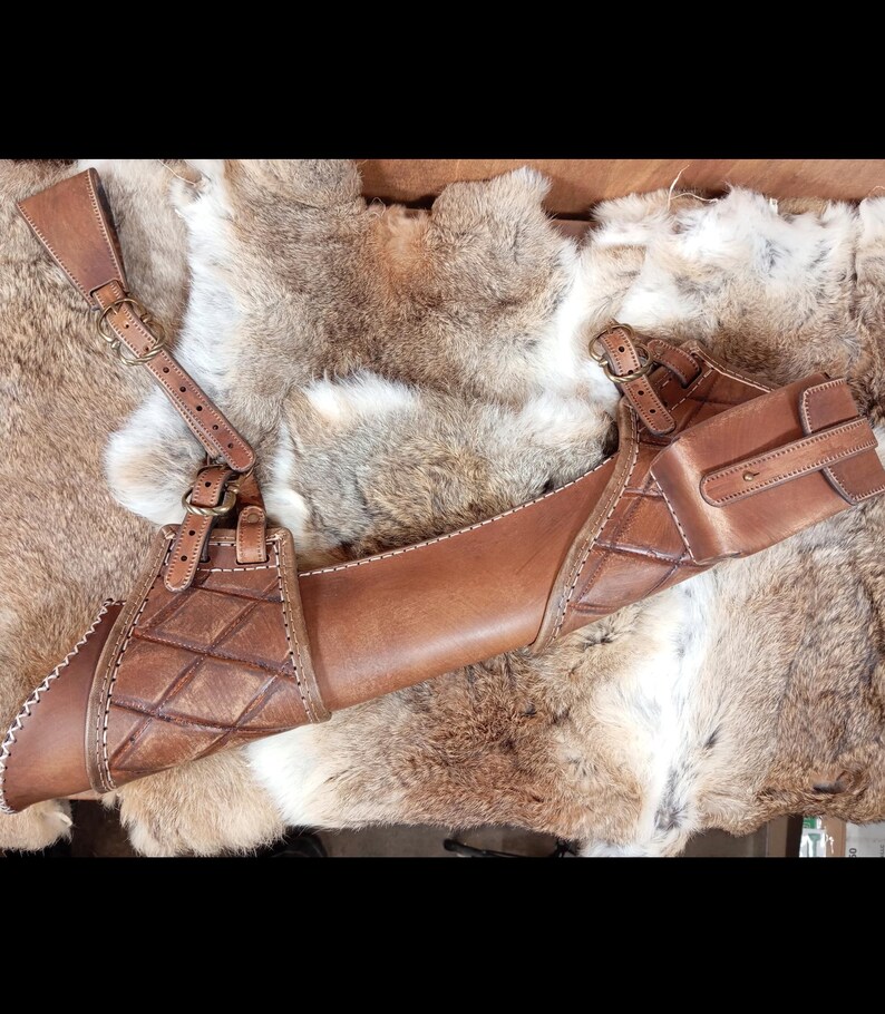 May include: A brown leather archery quiver with a shoulder strap and a small pouch. The quiver has a diamond pattern and white stitching. The leather is a medium brown color.