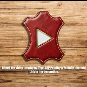 May include: A red leather hide-shaped patch featuring a central white triangle with a gold border. The text reads: Check the video tutorial on The LARP Peddler's Youtube channel. Link in the descrption.