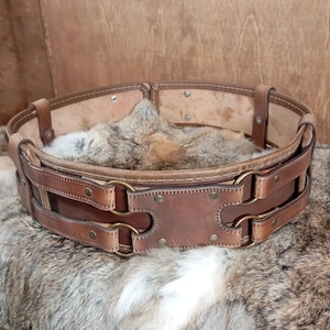 May include: A wide, brown leather belt with brass-colored metal rings and rivets. The belt has a double-layered design with a series of straps and loops. The belt is displayed on a furry, light brown surface.
