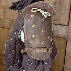 May include: Brown leather armour with metal studs and a strap. The armour is worn over a brown quilted garment.