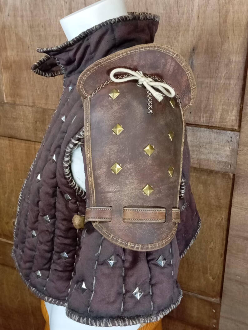 May include: Brown leather armour with metal studs and a strap. The armour is worn over a brown quilted garment.