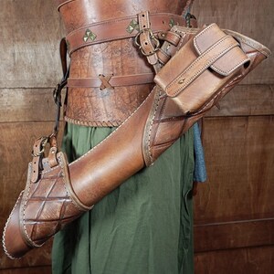 May include: A brown leather archery quiver with a matching arm guard and a small pouch. The quiver has a diamond pattern and is attached to a leather harness. The outfit is worn over green fabric.