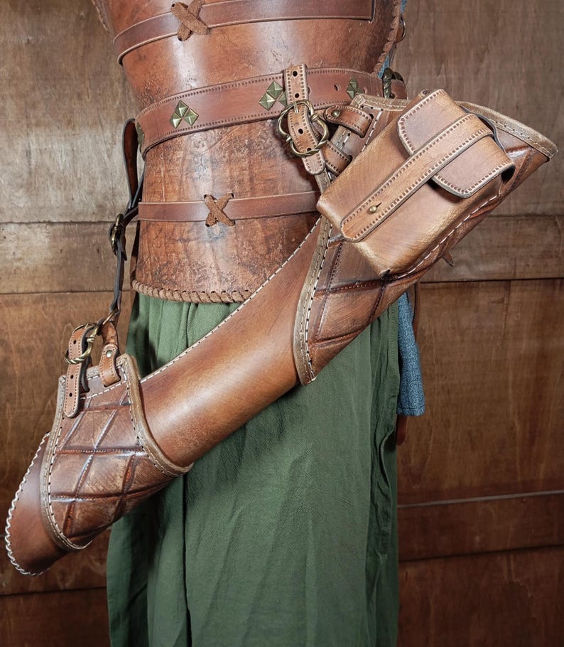 May include: A brown leather archery quiver with a matching arm guard and a small pouch. The quiver has a diamond pattern and is attached to a leather harness. The outfit is worn over green fabric.