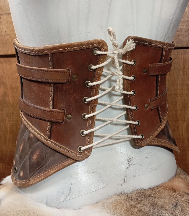 Puede incluir: Un cors&eacute; de cuero marr&oacute;n con cordones en la parte delantera y correas decorativas. El cors&eacute; presenta una secci&oacute;n inferior con dise&ntilde;o de diamantes y se muestra en un maniqu&iacute;. El cuero tiene un aspecto envejecido.