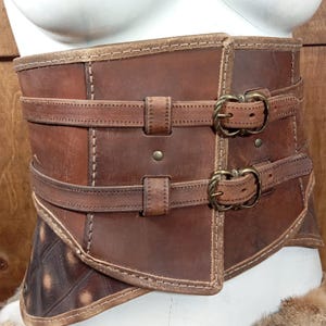 Puede incluir: Un cors&eacute; de cuero marr&oacute;n con dos correas y hebillas de lat&oacute;n. El cors&eacute; tiene un dise&ntilde;o cosido y se muestra en un maniqu&iacute; blanco. El cuero tiene un aspecto envejecido, con variaciones de color.