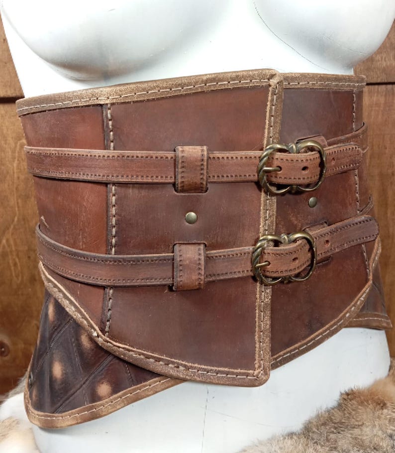 Puede incluir: Un cors&eacute; de cuero marr&oacute;n con dos correas y hebillas de lat&oacute;n. El cors&eacute; tiene un dise&ntilde;o cosido y se muestra en un maniqu&iacute; blanco. El cuero tiene un aspecto envejecido, con variaciones de color.