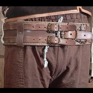 May include: A brown leather belt with multiple straps and brass buckles. The belt has a detailed stitched design and is worn with brown pants. The belt appears to be a part of a costume or historical garment.