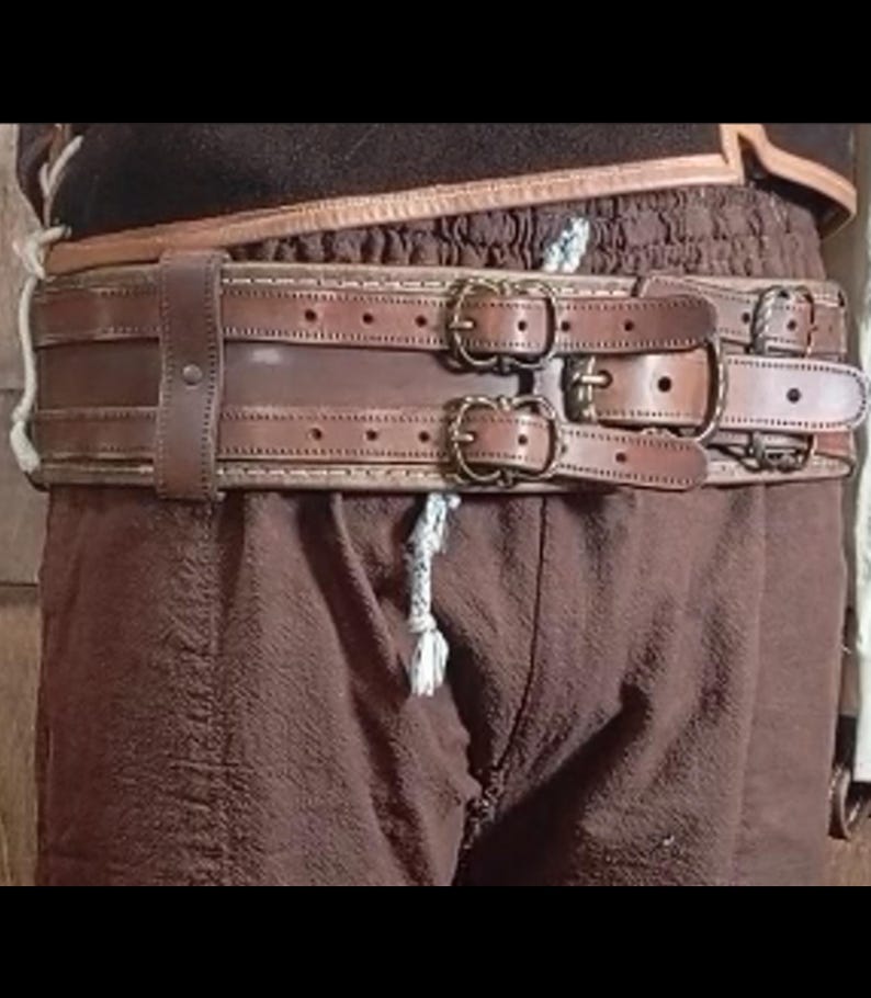 May include: A brown leather belt with multiple straps and brass buckles. The belt has a detailed stitched design and is worn with brown pants. The belt appears to be a part of a costume or historical garment.