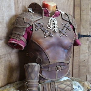 Puede incluir: Un peto y protectores de brazos de cuero marr&oacute;n con costuras decorativas y detalles met&aacute;licos. La armadura presenta un frente con cordones y se exhibe en un maniqu&iacute;. El interior est&aacute; forrado con tela roja.