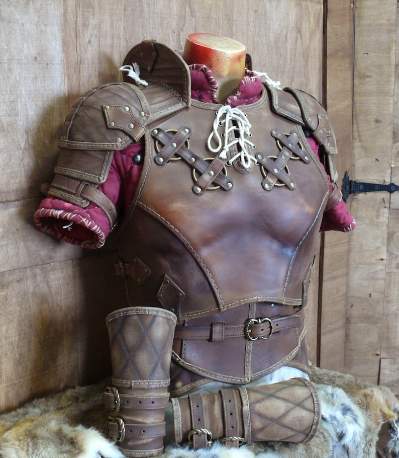 Puede incluir: Un peto y protectores de brazos de cuero marr&oacute;n con costuras decorativas y detalles met&aacute;licos. La armadura presenta un frente con cordones y se exhibe en un maniqu&iacute;. El interior est&aacute; forrado con tela roja.
