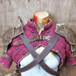 Puede incluir: Un chaleco de armadura medieval acolchado de color burdeos con detalles y correas de cuero marr&oacute;n. El chaleco presenta tachuelas met&aacute;licas y protectores de hombros. La armadura se exhibe en un maniqu&iacute;.