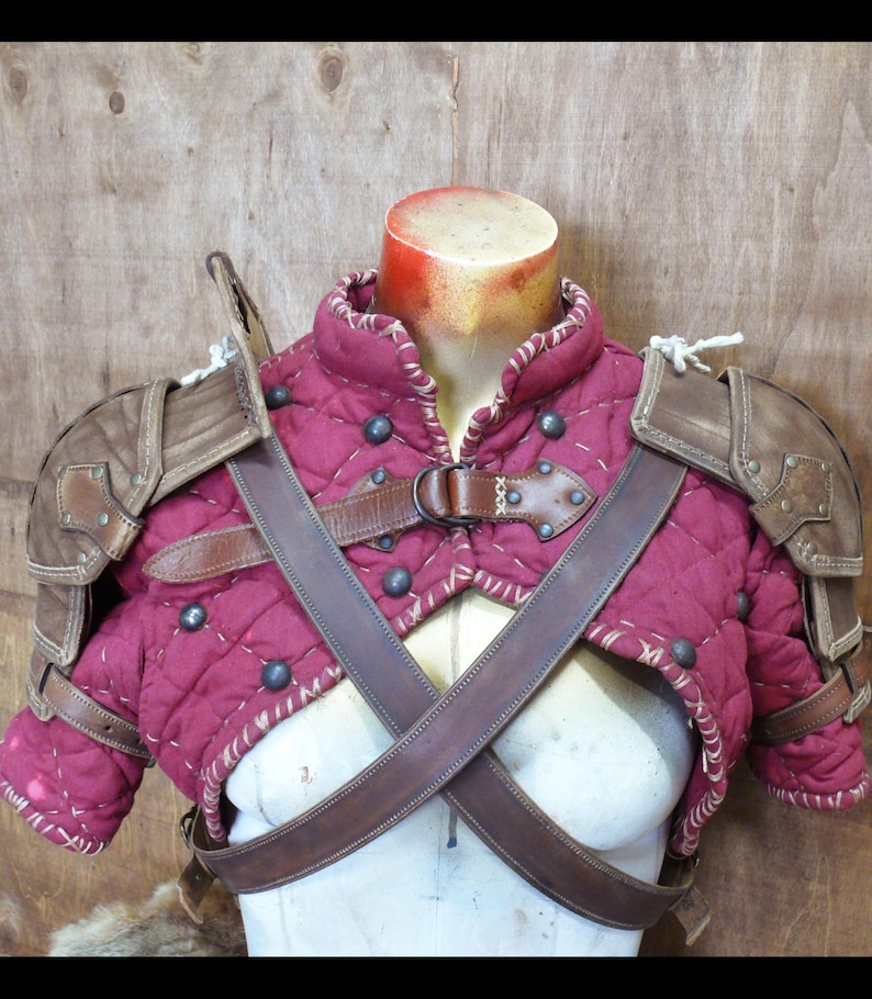 Puede incluir: Un chaleco de armadura medieval acolchado de color burdeos con detalles y correas de cuero marr&oacute;n. El chaleco presenta tachuelas met&aacute;licas y protectores de hombros. La armadura se exhibe en un maniqu&iacute;.