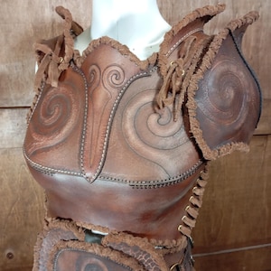 May include: A brown leather chest and shoulder armour piece featuring embossed spiral patterns, stitched edges, and frayed leather trim. The garment includes shoulder pauldrons, a central chest plate, and side lacing with metal eyelets for adjustment.