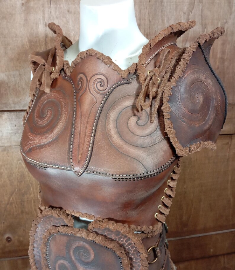 May include: A brown leather chest and shoulder armour piece featuring embossed spiral patterns, stitched edges, and frayed leather trim. The garment includes shoulder pauldrons, a central chest plate, and side lacing with metal eyelets for adjustment.