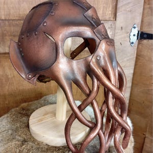 May include: A copper-colored helmet featuring a rounded dome with riveted panels and a face mask designed with multiple long, dangling tentacles extending downward. The structure is crafted with a metallic finish and visible seams throughout.