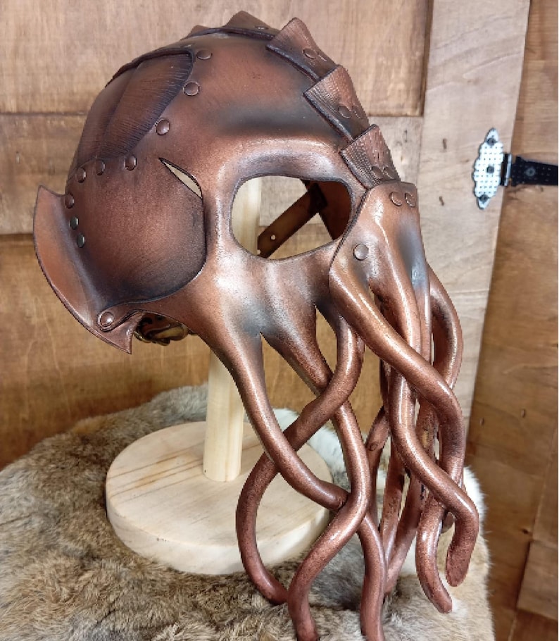 May include: A copper-colored helmet featuring a rounded dome with riveted panels and a face mask designed with multiple long, dangling tentacles extending downward. The structure is crafted with a metallic finish and visible seams throughout.