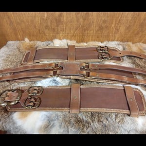 May include: Three brown leather belts with brass buckles and stitching. The belts are laid out on a fur-like surface. Each belt has a different design, with varying buckle styles and strap configurations.