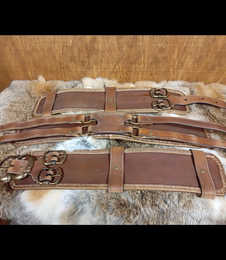 May include: Three brown leather belts with brass buckles and stitching. The belts are laid out on a fur-like surface. Each belt has a different design, with varying buckle styles and strap configurations.