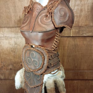 May include: A brown leather chest piece and belt set featuring embossed spiral and triskelion patterns. The armor includes shoulder guards, side lacing with leather cords, metal rings on the belt, and decorative frayed edges with faux fur accents.