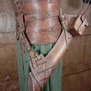 May include: A brown leather archery set, including a quiver, arm guard, and belt, with decorative stitching and metal accents. The set is worn over green fabric. The quiver is designed to hold arrows.