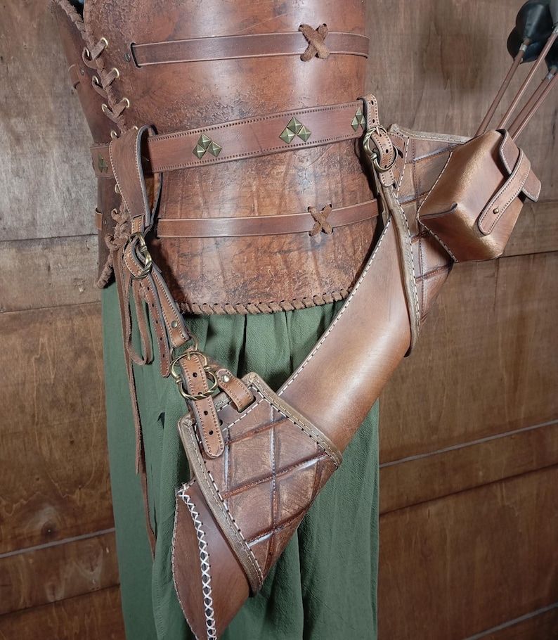May include: A brown leather archery set, including a quiver, arm guard, and belt, with decorative stitching and metal accents. The set is worn over green fabric. The quiver is designed to hold arrows.