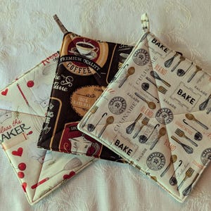May include: Three quilted pot holders with various kitchen-themed patterns. One features coffee cups and text, another has baking utensils and the word "BAKE", and the third has hearts and the text "Bless the BAKER".