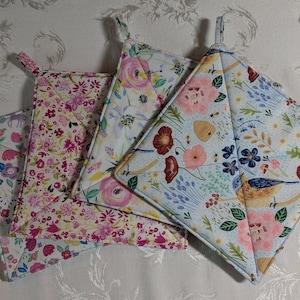 May include: Four quilted pot holders with floral patterns. The designs include pink and purple flowers, bees, and birds on a light blue background. Each pot holder has a loop for hanging.