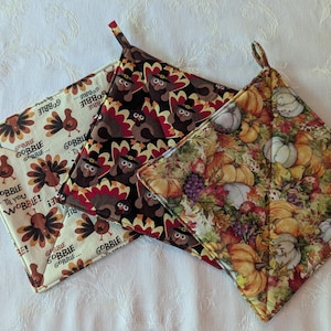 May include: Three quilted pot holders with loop handles. One features turkeys with pilgrim hats and the text "GOBBIE TIL YOU WOBBLE!". Another has a pumpkin and fall foliage pattern. The third has a turkey pattern.