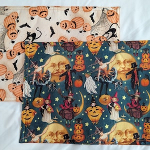May include: Two rectangular Halloween-themed placemats. One features orange pumpkins, black cats, and spiderwebs on a cream background. The other has a teal background with vintage Halloween characters, pumpkins, and moons. Both have a stitched border.