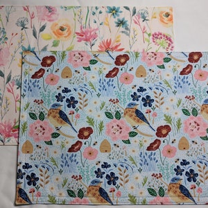 May include: Two floral patterned placemats. The top placemat is white with pink, yellow, and blue flowers. The bottom placemat is light blue with pink, red, and blue flowers, birds, and bees.