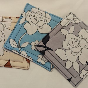 May include: Three square fabric coasters with floral designs. One is beige with brown accents, another is light blue, and the third is gray. Each coaster features a white rose and a black outline.