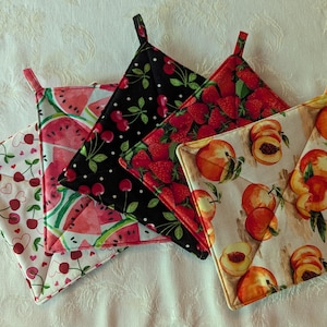 May include: A collection of five square pot holders with loop handles. Each pot holder features a different fruit pattern: cherries, watermelon, strawberries, peaches, and cherries on a black background. The pot holders are made of fabric.