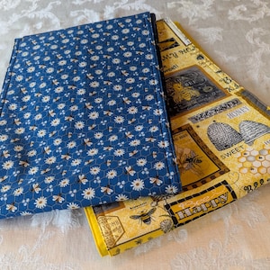 May include: Two fabric table runners, one blue with a white daisy and bee pattern and one yellow with a bee and honey themed print. The text on the yellow runner reads "Bee Happy" and "Bee Sweet".