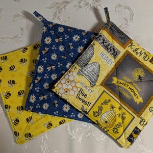 May include: Three square pot holders with bee and honey-themed patterns. One is yellow with bees, another is navy blue with bees and flowers, and the third is yellow with honey-related text and graphics. Each has a fabric loop.