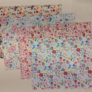 May include: Four floral patterned placemats in different colors. The placemats are rectangular and feature a variety of flowers and leaves in shades of pink, yellow, blue, and green.