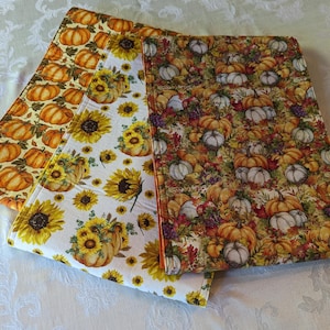 May include: Two fabric table runners with a fall theme. One runner features a white background with sunflowers and pumpkins. The other runner features a brown background with pumpkins, leaves, and grapes.