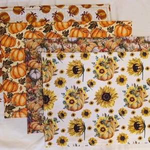 May include: Four placemats with a fall theme. The placemats feature a white background with a repeating pattern of sunflowers and pumpkins. The placemats are layered on top of each other, with the top placemat being the smallest and the bottom placemat being the largest.
