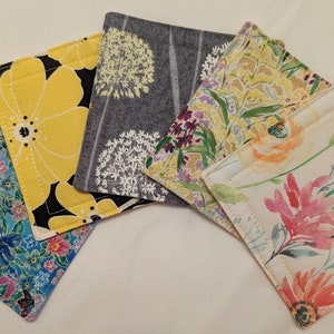 May include: Set of six fabric coasters with floral patterns. The coasters feature a variety of floral designs, including yellow flowers, purple flowers, and pink flowers. The coasters are all square and have a quilted texture.