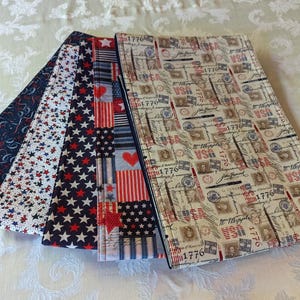May include: Four fabric placemats with patriotic designs. One placemat features a red, white, and blue stars and stripes pattern. Another placemat has a red, white, and blue pattern with hearts and stars. A third placemat has a blue and white pattern with red stars. The fourth placemat has a cream background with a red, white, and blue pattern of stamps and writing. The text on the placemat reads "1776".