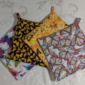 May include: A collection of four quilted pot holders with butterfly patterns. The pot holders feature various colors, including black with yellow butterflies, yellow with orange and black butterflies, and white with colorful butterflies. Each has a hanging loop.