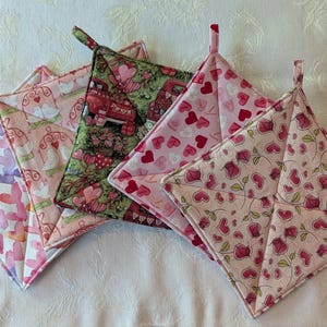 May include: A collection of five quilted pot holders in various heart-themed patterns. Designs include watercolor hearts, love birds, a red truck, and floral prints. Each pot holder has a loop for hanging.