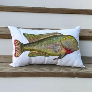 Puede incluir: Una almohada blanca con una ilustración colorida de un pez. El pez es verde, amarillo y rojo sobre un fondo blanco.