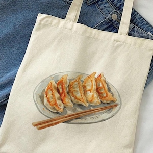 May include: A beige canvas tote bag with a design of a plate of gyoza and chopsticks. The bag has two long handles and the text "Olive Me Gifts" is printed on the bottom right corner.
