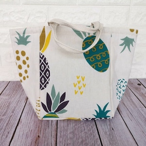 May include: A white canvas tote bag with a pineapple print. The bag has a green, yellow, and purple pineapple design. The bag has two white canvas straps.