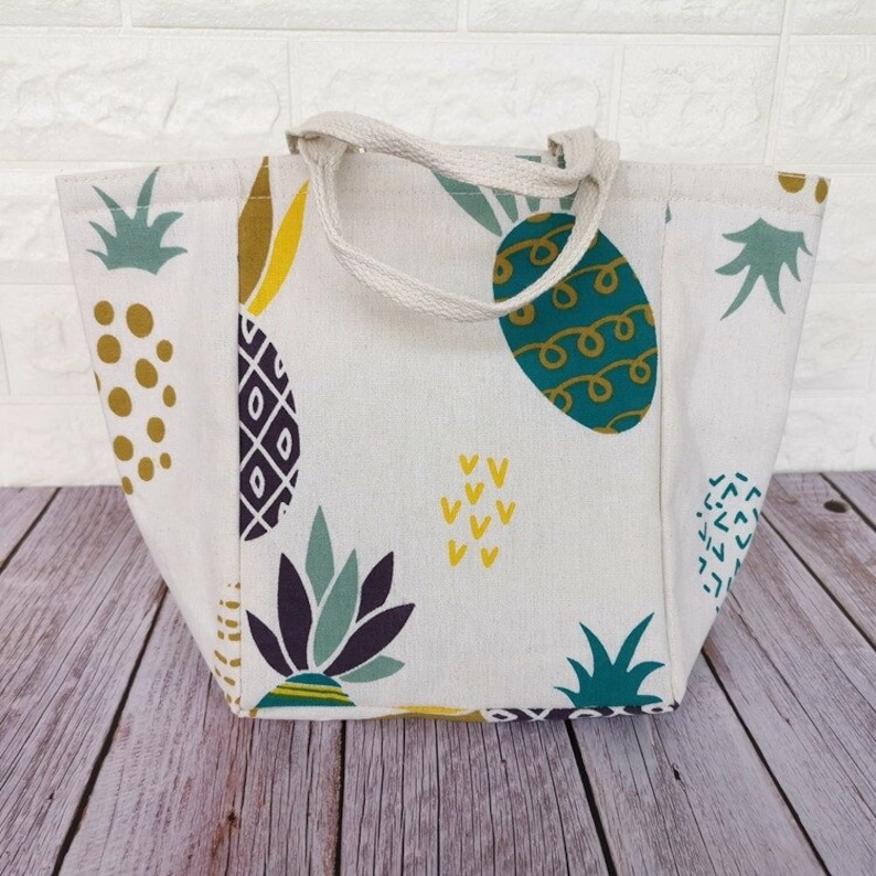 May include: A white canvas tote bag with a pineapple print. The bag has a green, yellow, and purple pineapple design. The bag has two white canvas straps.