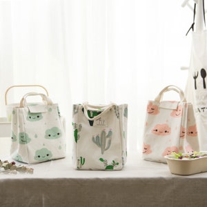 May include: Three white canvas lunch bags with different patterns. One bag has a cactus print, one has a cloud print with pink clouds, and one has a cloud print with green clouds.