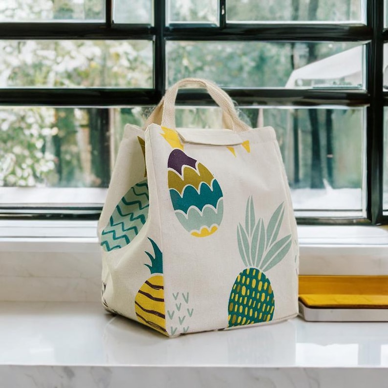 May include: A white canvas lunch bag with a colorful pineapple design. The bag has a zipper closure and two handles.