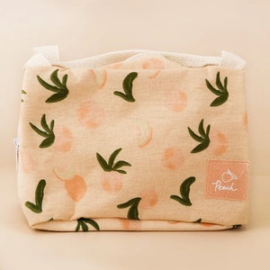 May include: A beige fabric lunch bag with a peach pattern and a white zipper. The bag features a peach and leaf design in shades of peach and green. A small pink label with the word "Peach" is sewn on the front.