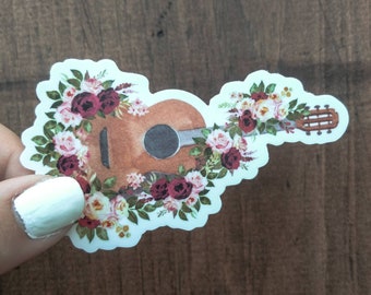 Op de afbeelding: Een sticker met een bruine akoestische gitaar met een bloemenontwerp. De gitaar is omringd door roze, witte en rode rozen en groene bladeren.